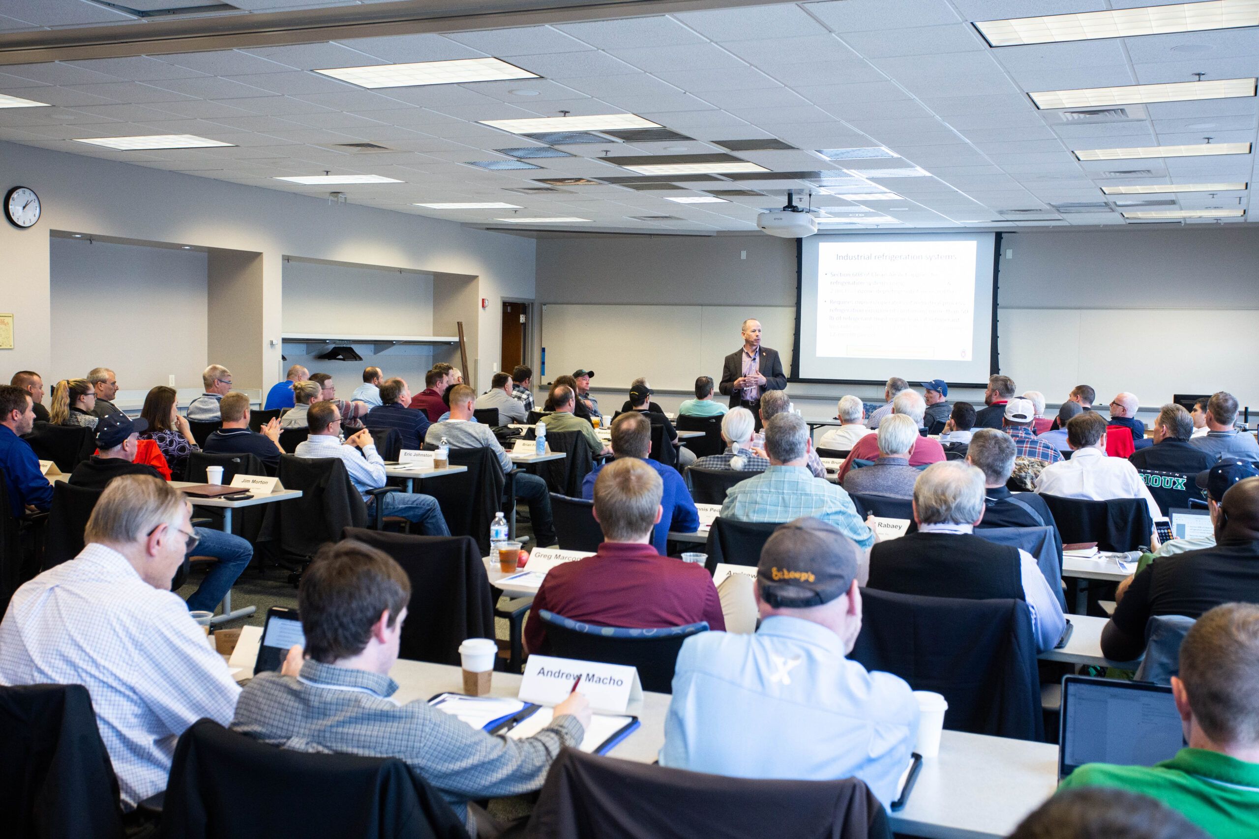 A man delivers a presentation to a large group of engaged adult learners seated in a classroom.